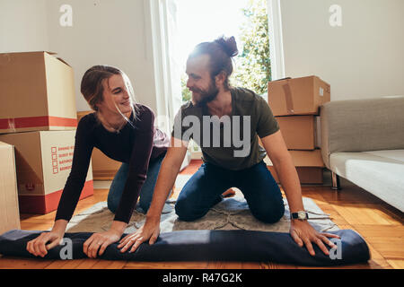 Smiling man and woman sitting on a carpet and rolling it for packing. Couple looking at each other while packing their household items. Stock Photo