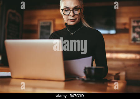 Senior businesswoman reading some documents at coffee shop. Female in casuals sitting at cafe table with laptop reading business report. Stock Photo