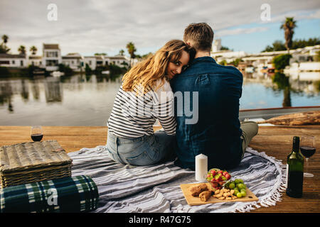 Rear view of a couple in love sitting side by side beside a lake. Couple on a day out sitting together with picnic food and drinks facing the lake wit Stock Photo