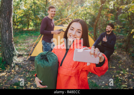 Picture of beautiful young woman takes selfie and smiles. She holds sleeping bag. Young men on back poses as well. They work with tent.