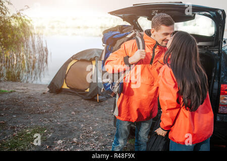 Lovely picture of young couple kissing. They wear orange jackets. People standing close to each other. He has backpack. Couple is at opened trunk and tent. Stock Photo