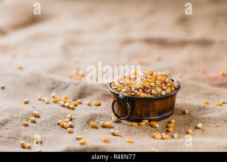 Corn in a little pot lying on a brown, structured background, sprinkled with more corn Stock Photo