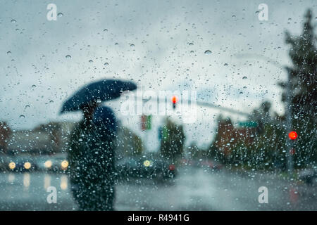 Blurred people silhouettes crossing a street, holding an umbrella on a rainy day; selective focus on the drops of rain on the windshield; California Stock Photo