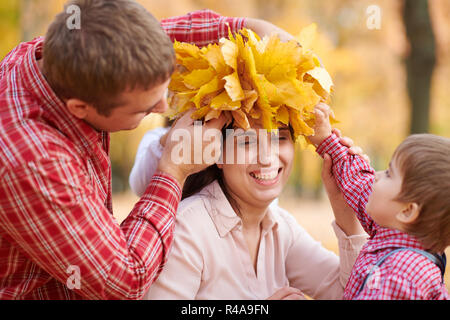 Father and son put yellow fallen leaves on mother head. Happy family is in autumn city park. Children and parents. They posing, smiling, playing and h Stock Photo
