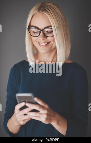 Smiling young businesswoman sending a text message on her cellphone while standing against a gray background Stock Photo