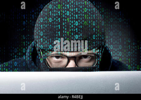 binary code on the background of a hacker in a balaclava and glasses looks out from behind a laptop and looks into the camera Stock Photo
