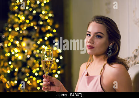 Portrait of a beautiful young woman is sitting in a dark chic hall with a Christmas tree dressed up in a garland in ampir chair with a glass of champa Stock Photo