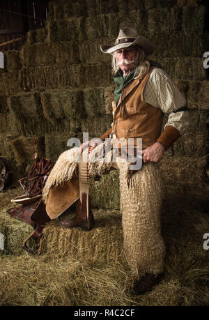Authentic old west cowboy portrait with wooly chaps, pistol, cowboy hat, leather vest, bandanna in stable Stock Photo