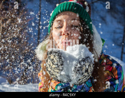 Beauty Winter Girl Blowing Snow in frosty winter Park. Outdoors. Flying Snowflakes. Sunny day. Backlit. Joyful Beauty young woman Having Fun in Winter Park. Stock Photo