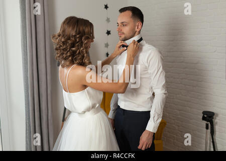 the bride and groom in the morning at the hotel on a wedding day. Dressing a newly-married couple Woman help fixing bowtie to groom. Stock Photo