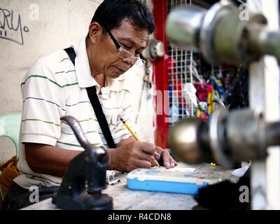 ANTIPOLO CITY, PHILIPPINES - NOVEMBER 24, 2018: A locksmith and rubber stamp maker works on his makeshift shop at a sidewalk along a busy street. Stock Photo