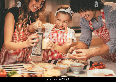 Happy parents and their daughter are preparing meal together in the kitchen. Little girl and her mother are putting cheese on the pizza and her father Stock Photo