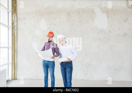 male engineers, architects in helmets stand by the window and hold a roll of paper with a building drawing Stock Photo