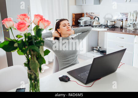 Woman relaxing after working on laptop on modern kitchen. Young tired freelancer resting with arms raised and eyes closed Stock Photo