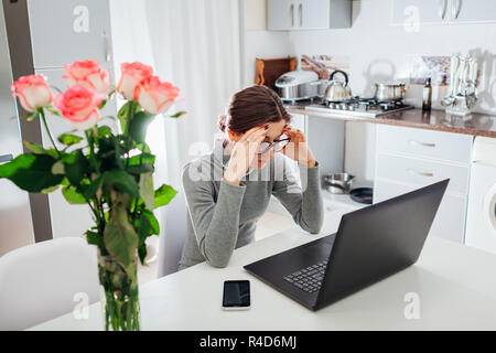 Woman working on laptop on modern kitchen. Young tired freelancer having headache. Lady upset Stock Photo