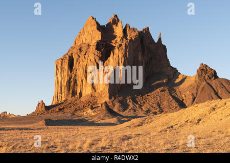 The dramatic rock formation of Shiprock rises above a grassy landscape in the early morning light of golden hour Stock Photo