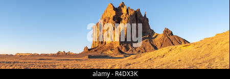 Panorama of the Shiprock rock formation rising above a grassy landscape in the early morning light of golden hour Stock Photo