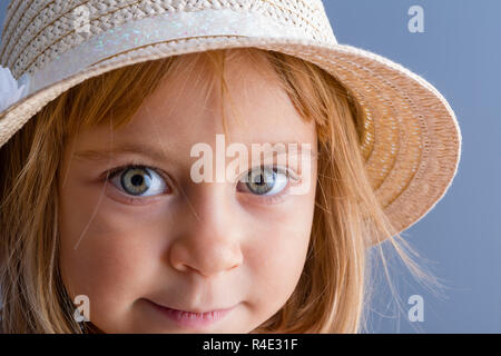 Pretty little girl with large green eyes wearing a trendy straw hat looking at the camera with a quiet smile
