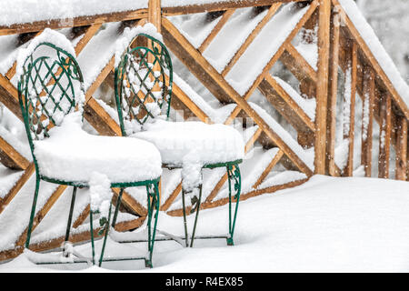 Closeup of two cast iron chairs covered in snow on home, house wooden deck, railing, fence in winter, snowing, falling snowflakes, snowstorm, storm in Stock Photo