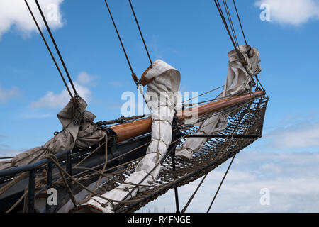 bowsprit and jib boom with reefed sails on the bow of a historic sailing ship against a blue sky with clouds Stock Photo