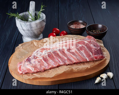 Raw pork ribs on crumpled paper on a bamboo cutting board, with rosemary twigs, tomatoes, garlic cloves, peppercorns and coarse pink salt Stock Photo