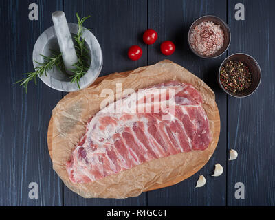 Raw pork ribs on crumpled paper on a bamboo cutting board, with rosemary twigs, tomatoes, garlic cloves, peppercorns and coarse pink salt Stock Photo