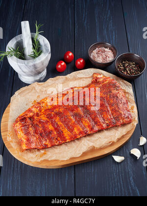 Marinated raw pork spare ribs on crumpled paper on a bamboo cutting board, with rosemary twigs, tomatoes, garlic cloves, pepper corns and coarse pink  Stock Photo