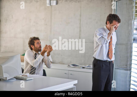 Mid-adult business man crying and rubbing his eyes while his colleague gestures to him in an office. Stock Photo