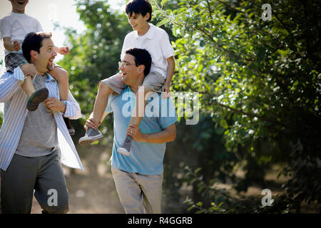 Two smiling mid-adult men carrying their young sons on their shoulders through an apple orchard in the sunshine. Stock Photo