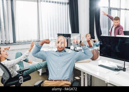happy young african american man showing biceps and smiling at camera while colleagues having fun behind in office Stock Photo