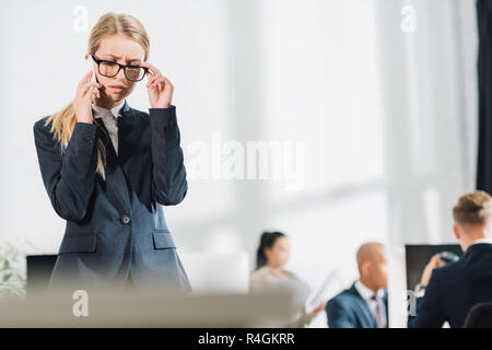 focused young businesswoman talking by smartphone and adjusting eyeglasses in office Stock Photo