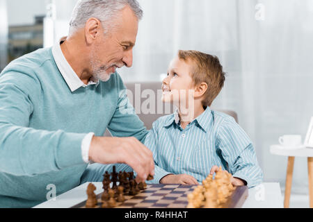 side view of happy grandson and grandfather looking at each other while playing chess at home Stock Photo
