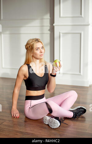 Young slim woman with an athletic body long blonde hair, dressed in a pink sports top and leggings, sits in a bright yoga room with a large panoramic  Stock Photo