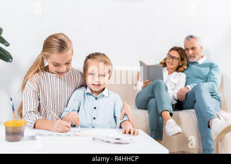 sister and brother painting at table while their grandparents sitting on sofa behind at home Stock Photo