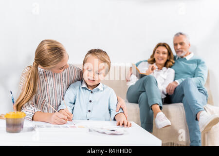 child helping little brother to painting at table while their grandparents sitting on sofa behind at home Stock Photo