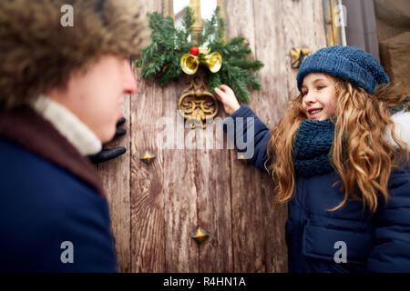 Little girl helps father and hanging Christmas wreath on the door. Cute curly daughter spend time with parents on holidays. New Year home decorations and preparations. Snowy winter outdoors. Stock Photo