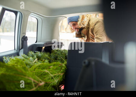 Bearded man loading christmas tree into the trunk of his car, inside view. Hipster puts fir tree into the back of his hatchback. Convertible auto interior with practical folding seats for boot space. Stock Photo