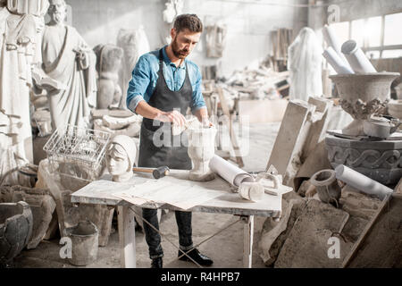 Man in blue t-shirt and apron working in the old atmospheric studio brushing stone vase on the table Stock Photo