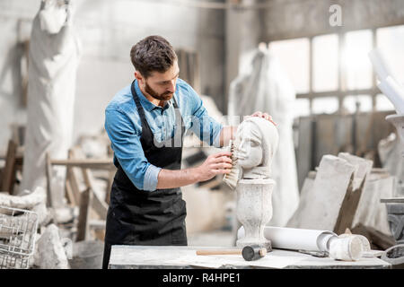 Handsome sculptor brushing stone head sculpture on the table in the atmospheric studio Stock Photo