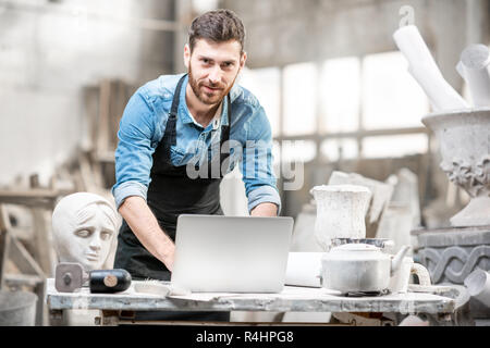 Handsome sculptor working with laptop at the working place in the old studio with sculptures on the background Stock Photo