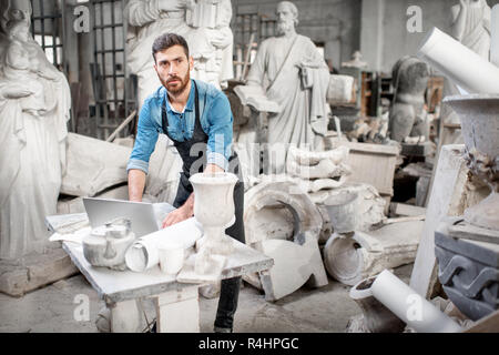 Handsome sculptor working with laptop at the working place in the old studio with sculptures on the background Stock Photo