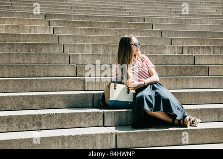 Beautiful tourist girl or student in sunglasses and with a backpack sits on the stairs and rests. Lifestyle Stock Photo