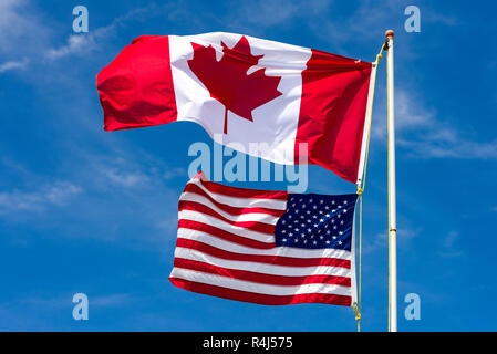 Large Canadian flag above smaller American flag on a white pole in the wind against a blue sky Stock Photo