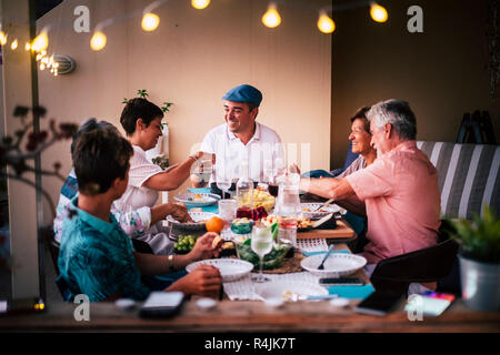 Dinner time in friendship with different ages people all together having fun and enjoying the night with smiles and happiness. Mobile phones defocused Stock Photo
