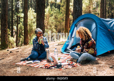 two different ages caucasian woman in free camping together in the pines forest eating a sandwich - freedom and independence for adult people - altern Stock Photo