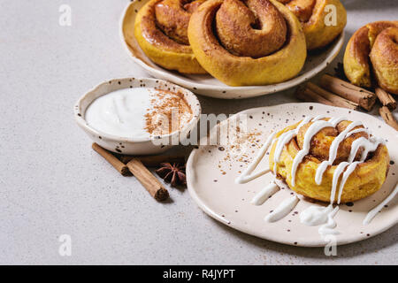 Homemade pumpkin cinnamon bun rolls sweet autumn baked dessert with cream cheese sauce in spotted ceramic plates with cinnamon sticks over white marbl Stock Photo