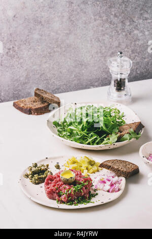 Beef tartare with quail egg in shell, rye bread, cutting pickled cucumbers, capers, red onion, chives and arugula salad served in spotted ceramic plat Stock Photo