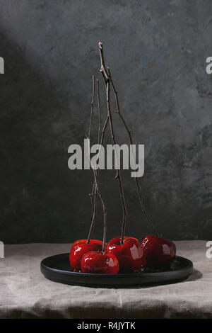 Red caramel apples sweet autumn or Christmas dessert served with branches in black ceramic plate on linen table cloth with grey wall at background. Da Stock Photo
