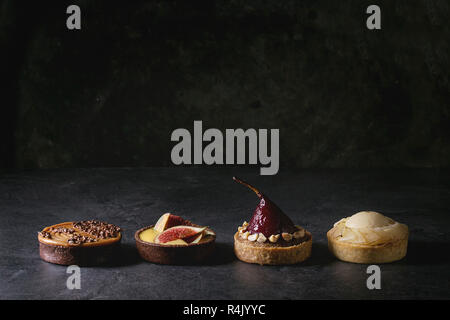 Variety of sweet tartlets with chocolate, caramel, pears, figs in row on black texture table. Stock Photo