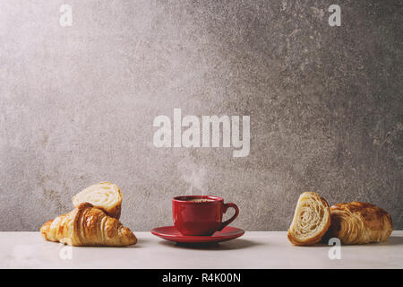 Fresh baked whole and sliced croissant with red cup of coffee espresso on white marble table with grey wall at background. Caffe breakfast Stock Photo